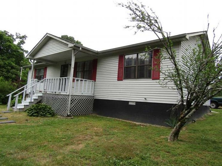 White, single-story house with a front porch, white siding, and red shutters, set on a grassy yard with a small tree nearby.