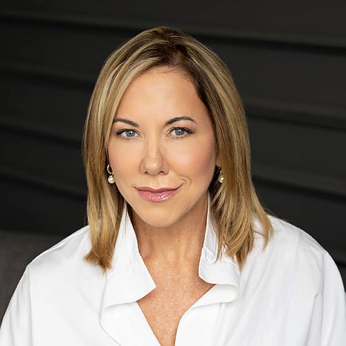 Head-and-shoulders portrait of a smiling blonde woman in a white blouse against a dark background.
