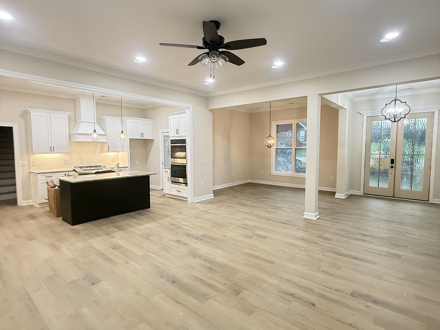 Open-concept kitchen with white cabinets, dark island, pendant lights, and double ovens beside a spacious living area.