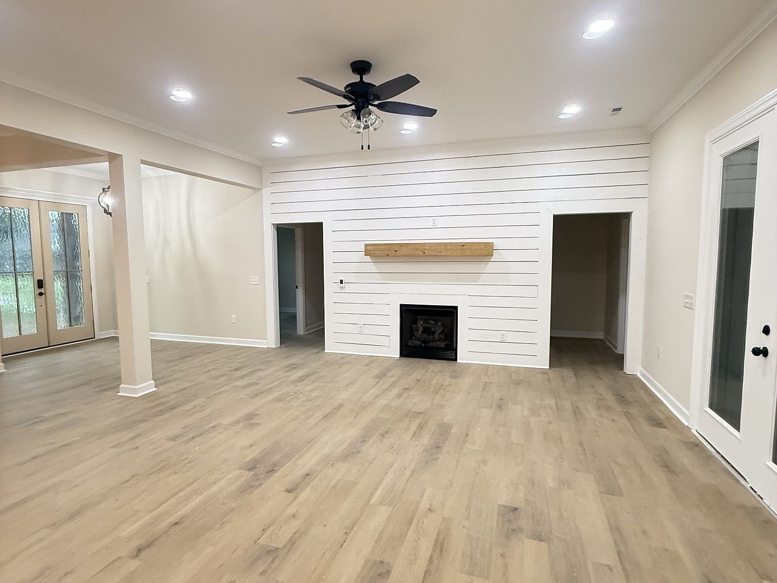 Living room with a white shiplap feature wall around a fireplace and a wooden mantle, ceiling fan, and recessed lights. Far left: French doors to outdoors.
