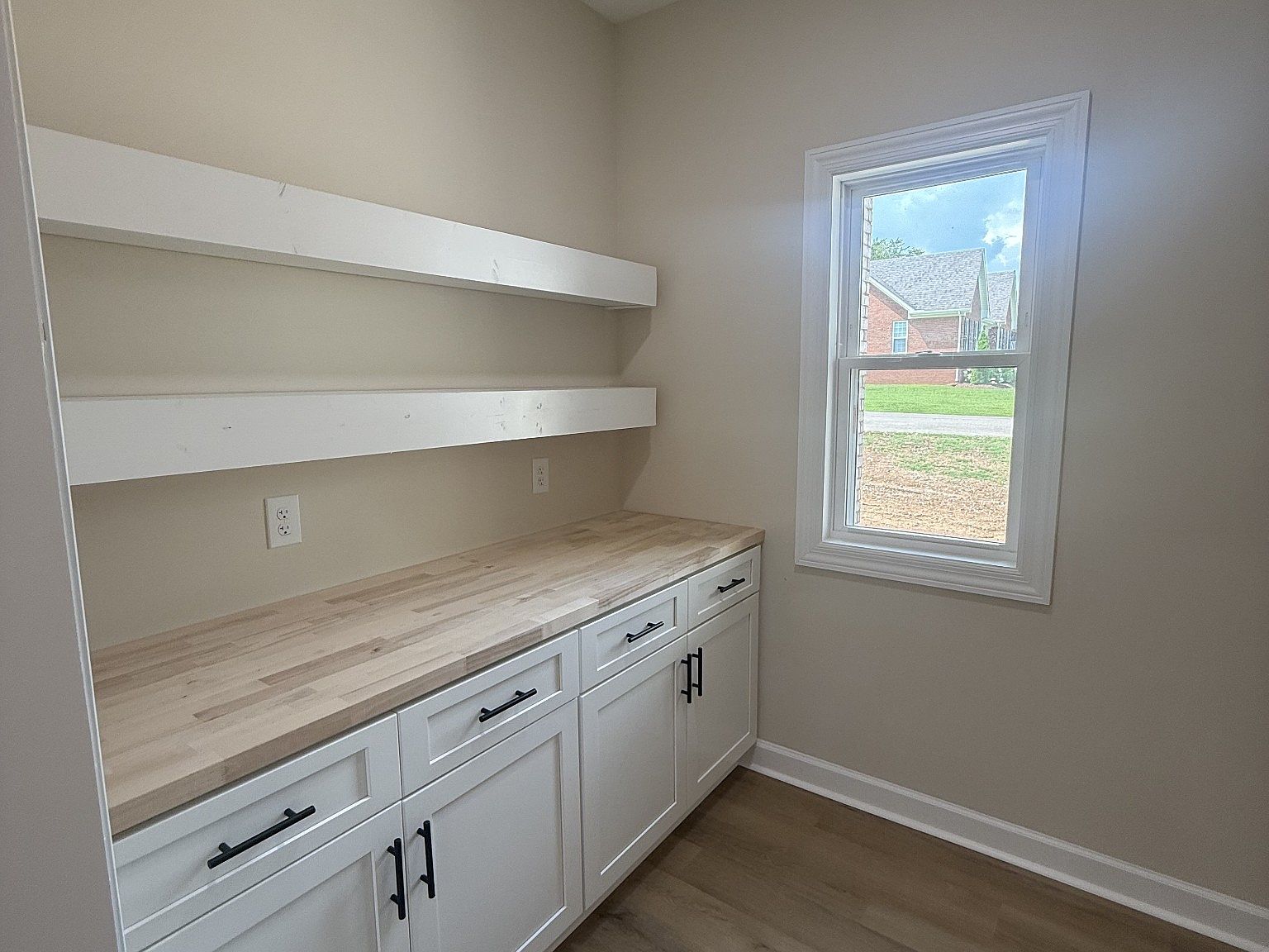 Light-filled utility room with white cabinets, a butcher-block counter, two long wall shelves, and a window showing a suburban yard