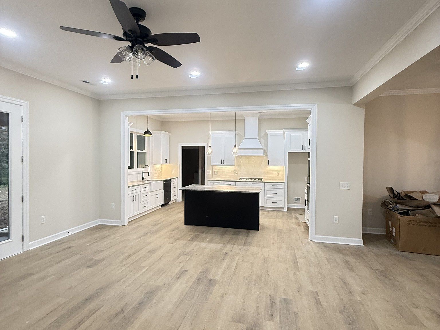 Open-concept kitchen with white cabinets, a black island, pendant lights, and a stainless range hood; light wood floors.