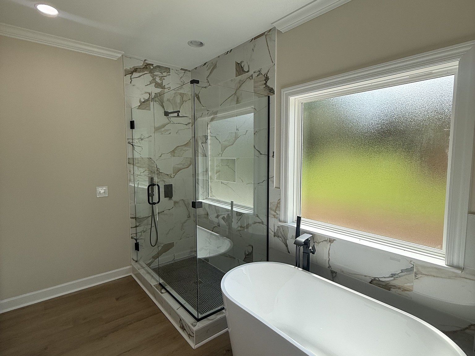 Modern bathroom with a glass-enclosed shower featuring black hardware and white marble tile, adjacent to a frosted window and a white freestanding oval tub.