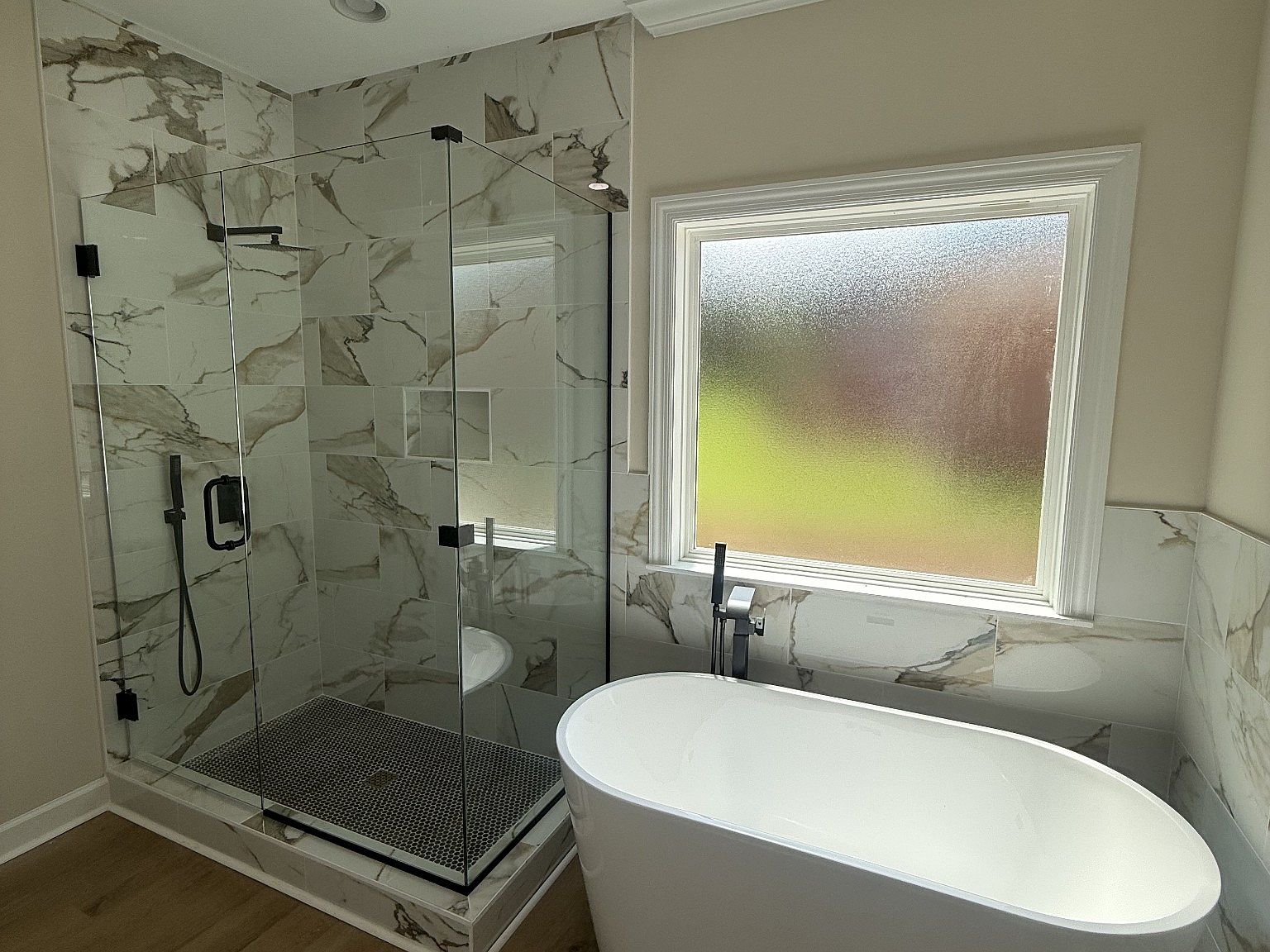 Modern bathroom with a glass-walled walk-in shower featuring marble-pattern walls, black fixtures, and a freestanding white tub by a frosted window.