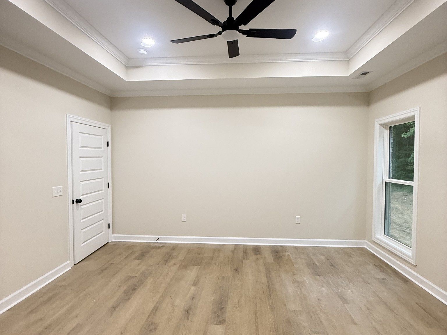 Empty beige room with white trim, a white door on the left, a window on the right, a ceiling fan, and light wood flooring.