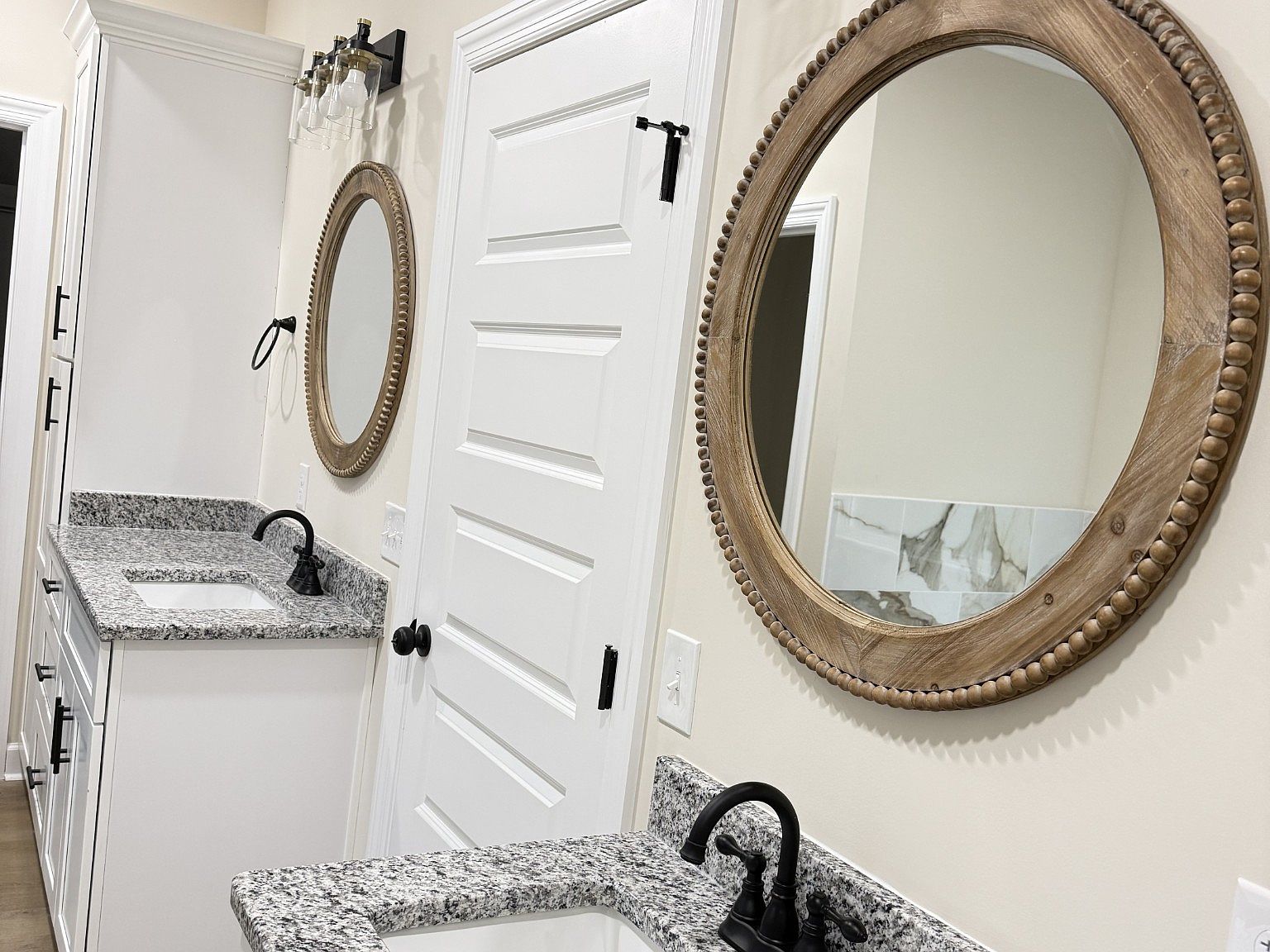 Double vanity with granite countertops, black faucets, white cabinets, and round beaded-wood mirrors above each sink in a bright bathroom.