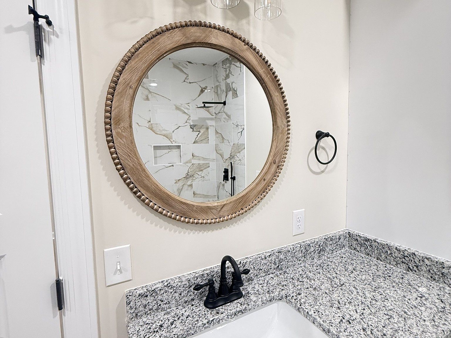 Round wooden mirror with beaded edge above a speckled granite bathroom counter and black faucet.