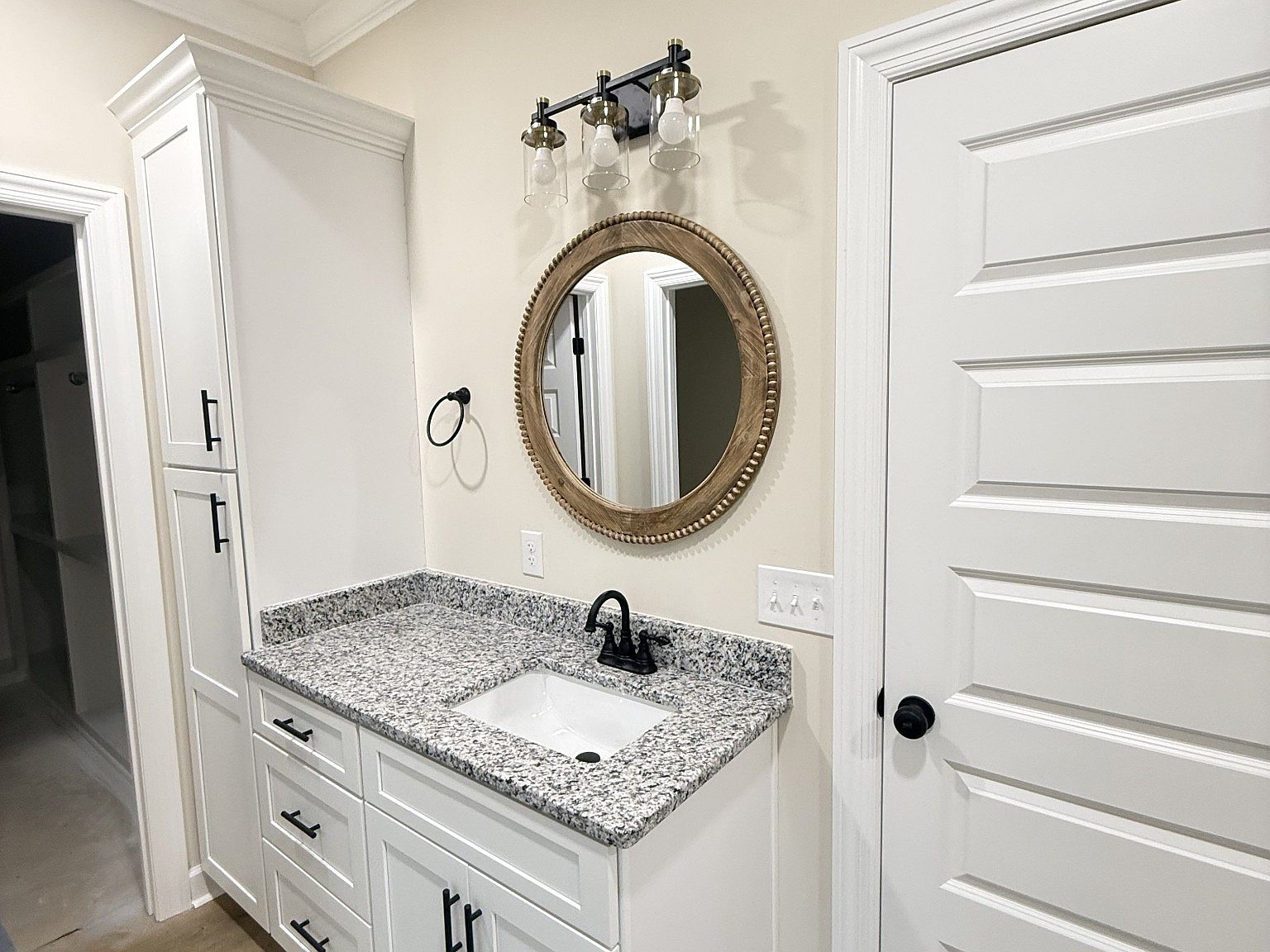 Bathroom vanity with granite countertop, white cabinets, a round beaded-wood mirror, black faucet, and a 4-light glass fixture above it