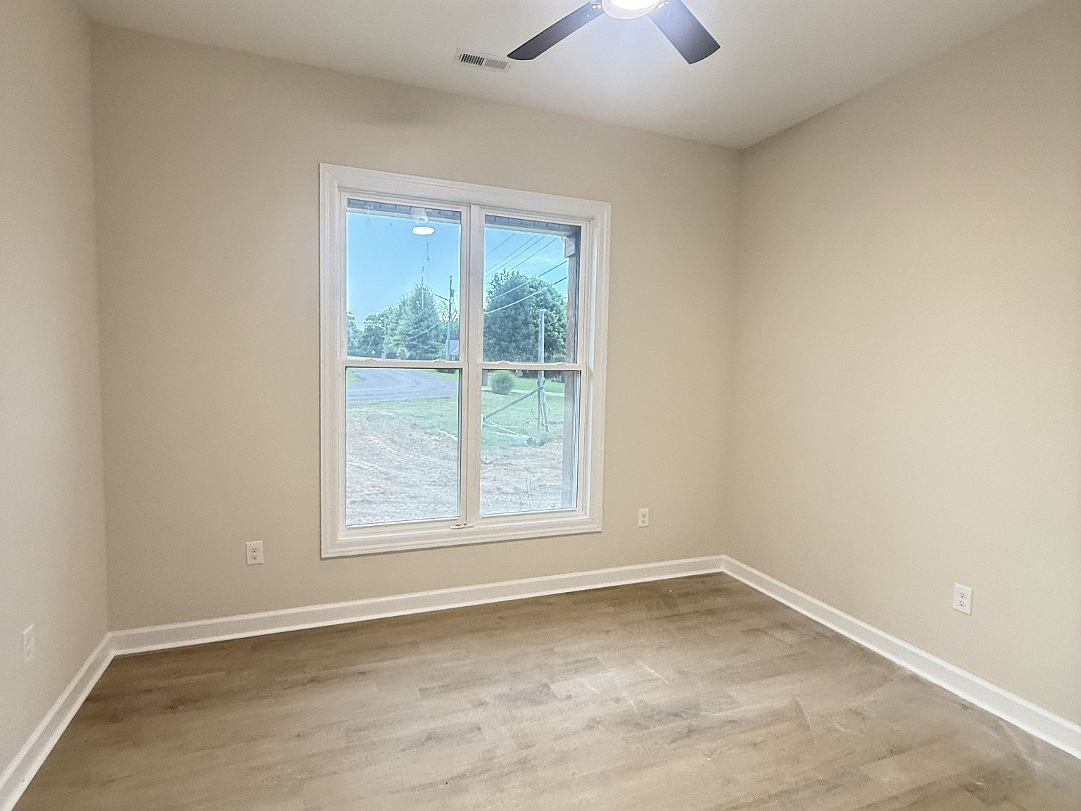 Unoccupied beige bedroom with a large window, light wood floor, and a ceiling fan, empty and ready for furnishing.