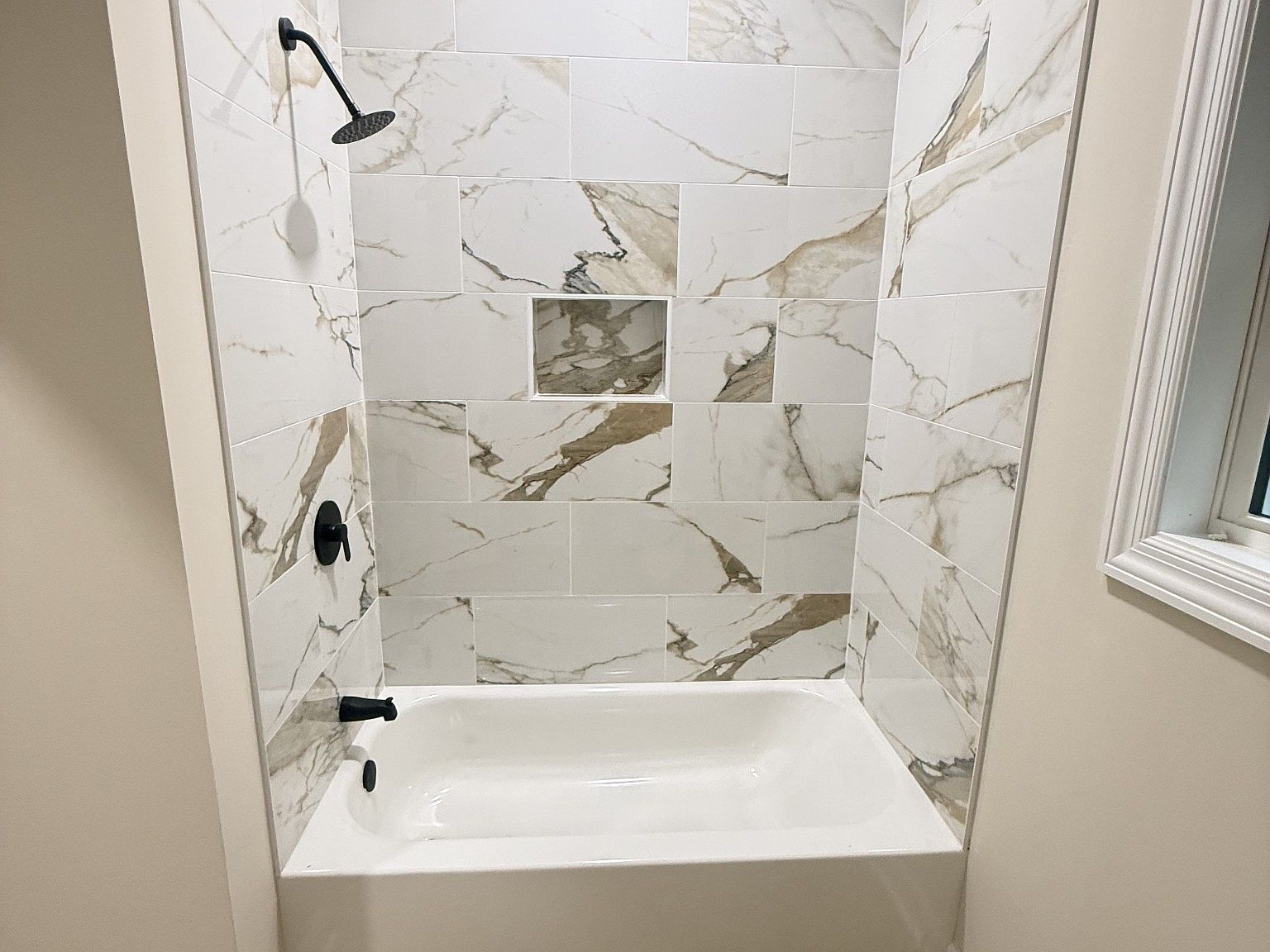 Shower/tub combo with white marble tile and a recessed niche built into the wall. (black fixtures)