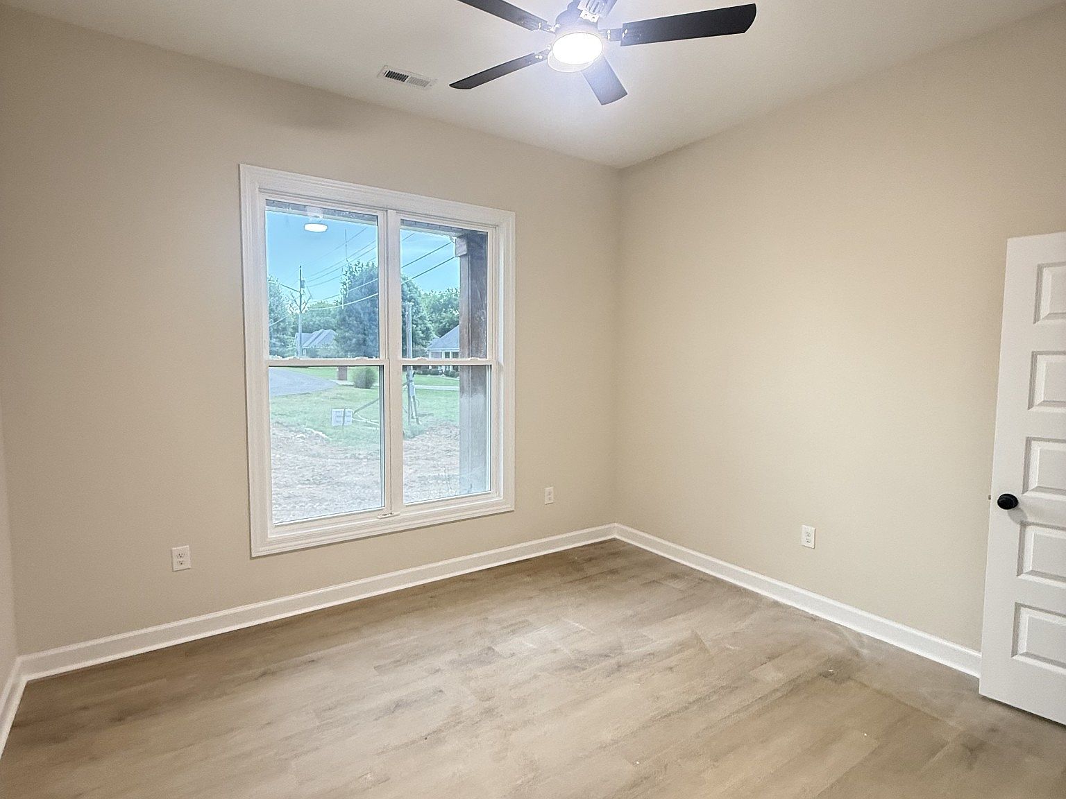 Empty beige bedroom with white trim, large double-hung window, light wood floors, and a ceiling fan overhead