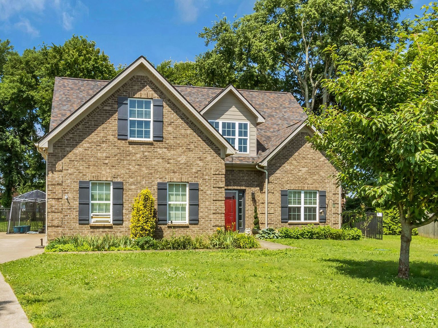 110 Bancroft Ct photo 3 Brick suburban house with blue shutters, a red front door, and a well-kept green lawn in front of a bright blue sky.