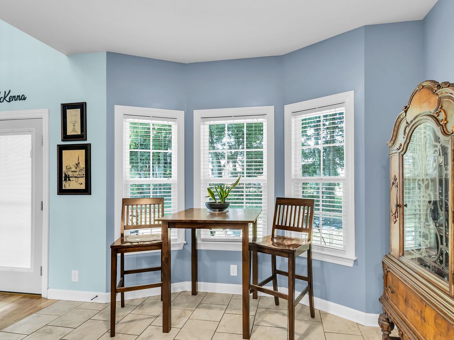 110 Bancroft Ct photo 13 Breakfast nook with a wooden table and two chairs by three white-framed windows, blue walls, and a potted plant centerpiece, sunlit space.