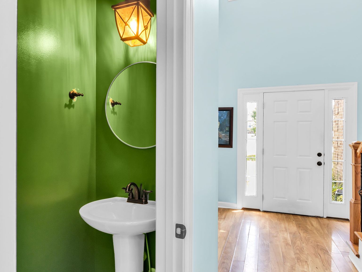 110 Bancroft Ct photo 18 Bright entryway toward a lime-green bathroom with pedestal sink, brass faucet, and round mirror; blue hallway with a white front door and sidelights.