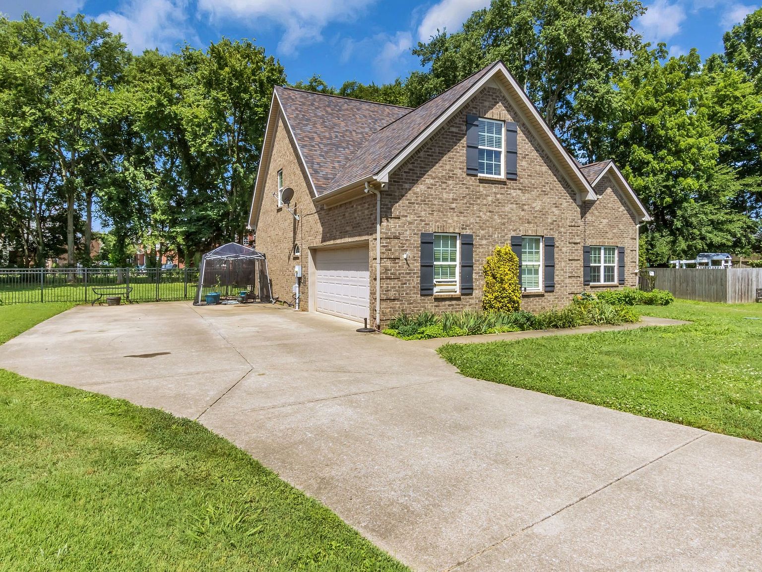 110 Bancroft Ct photo 34 Brick suburban house with a two‑car garage, curved concrete driveway, and green lawn framed by tall trees; a small greenhouse sits near the driveway on the left.