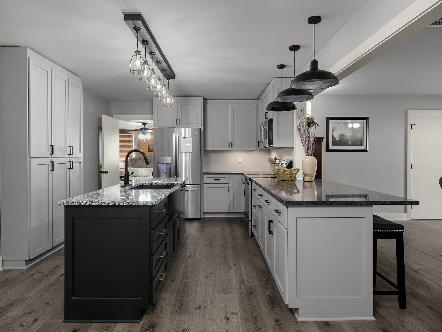 Modern kitchen with grey cabinets, granite island, and stainless steel appliances; pendant lights over the island and a walk‑through to a living area in the background