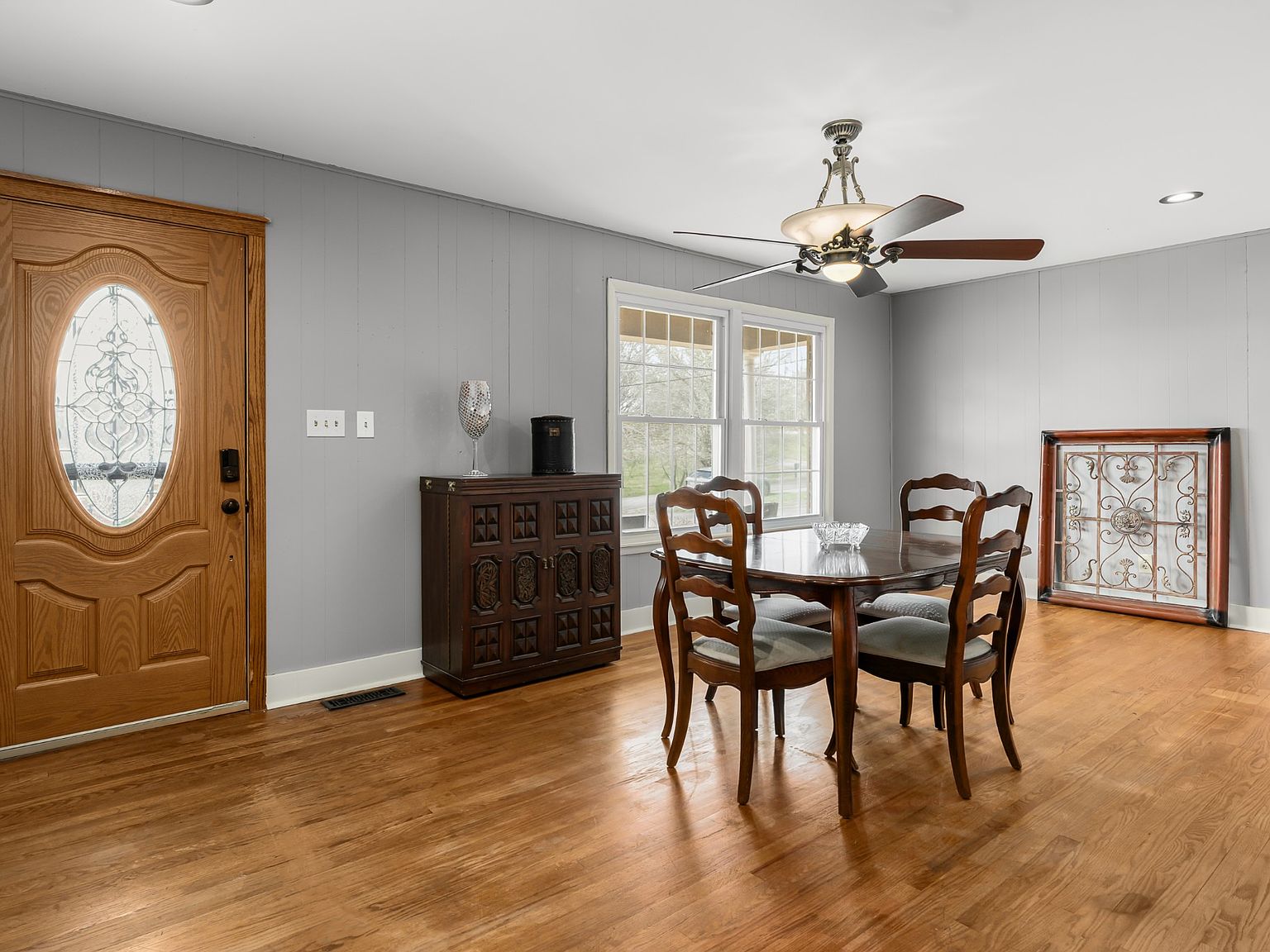 Dining room with a round wooden table and four carved chairs, gray paneled walls, and a ceiling fan above.