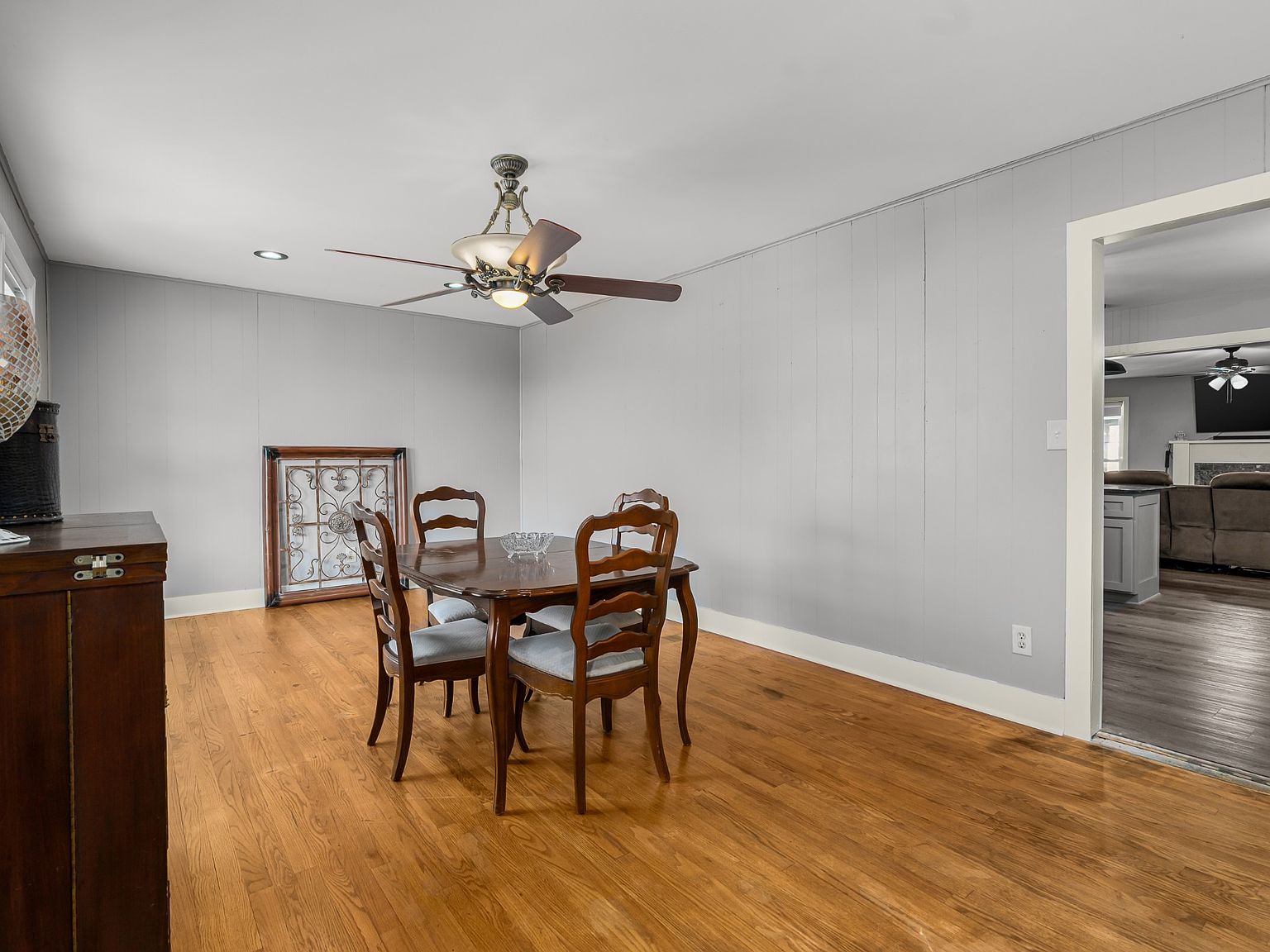 Wood dining table with four carved chairs in a light gray wainscoted dining room and hardwood floors, ceiling fan above.