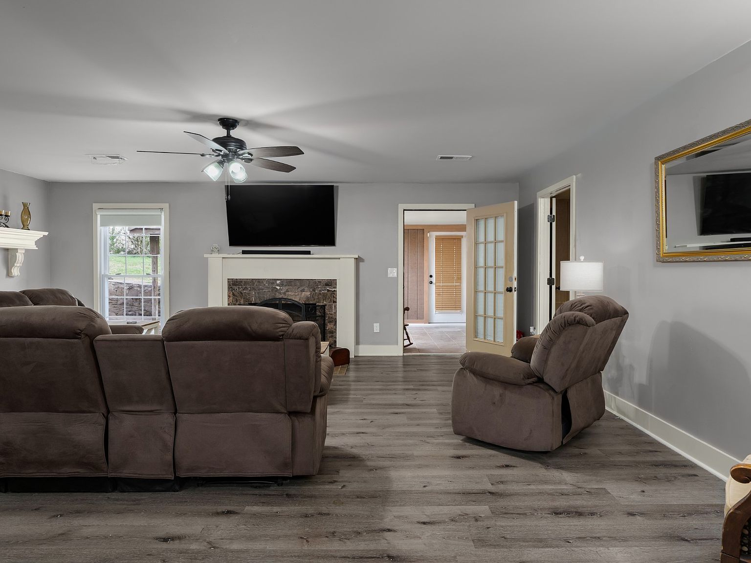 Living room with a brown sectional, a recliner, and a fireplace with a mounted TV above it; ceiling fan overhead and French doors to the outside.