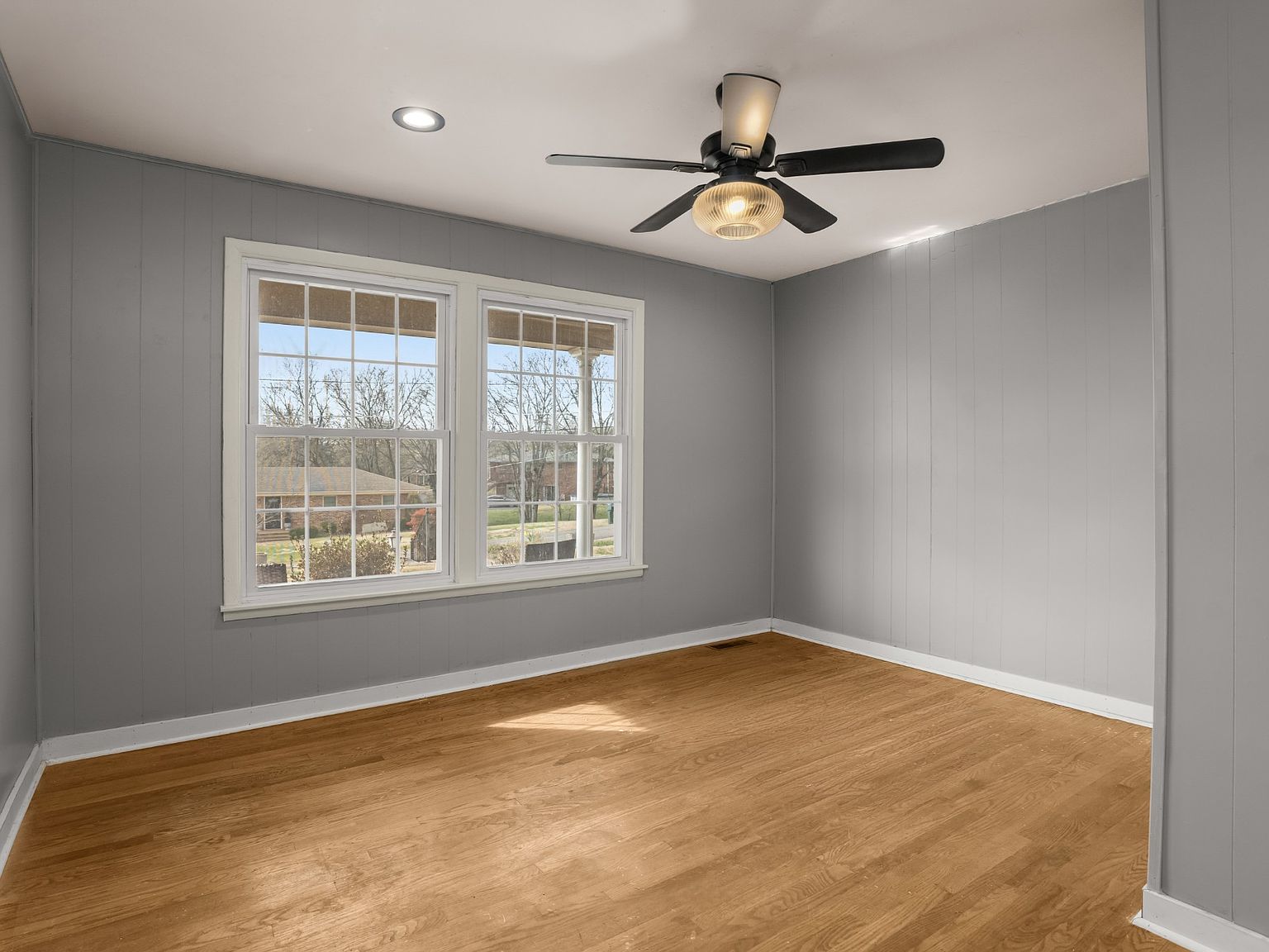 Empty room with gray paneled walls, a wooden floor, and a large double window letting in daylight, plus a ceiling fan.