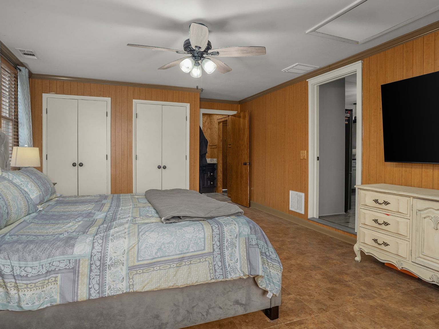 Bedroom with wood-paneled walls, queen bed in foreground, ceiling fan, and a dresser with a mounted TV on the wall.