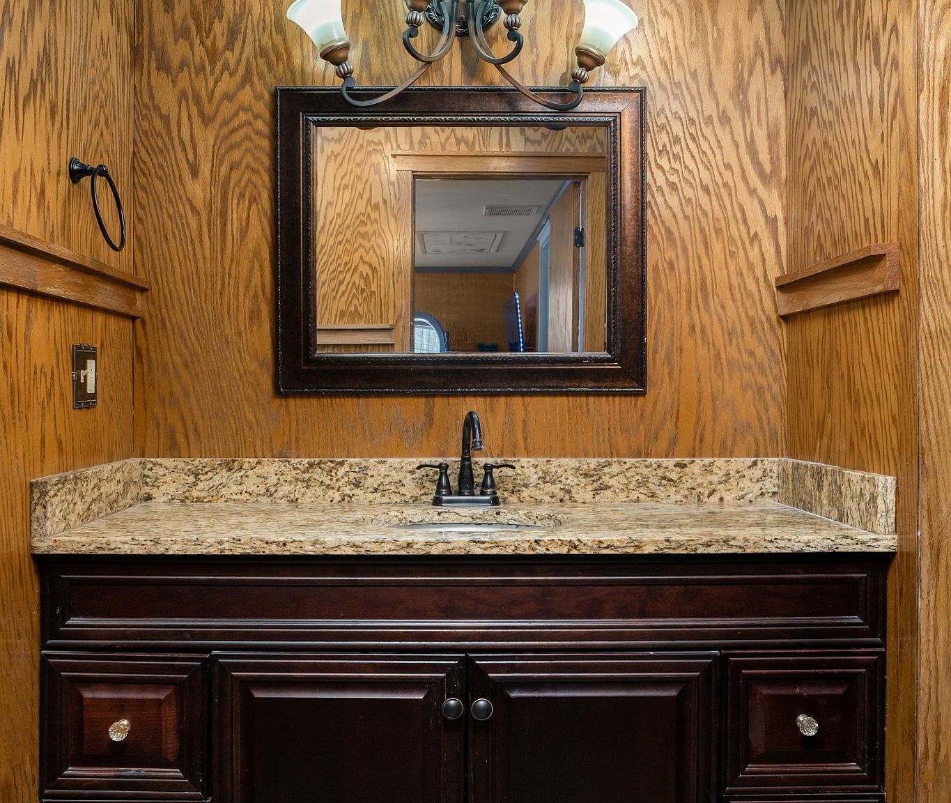 Traditional bathroom vanity with dark wood cabinets, beige granite countertop, and a framed mirror above.