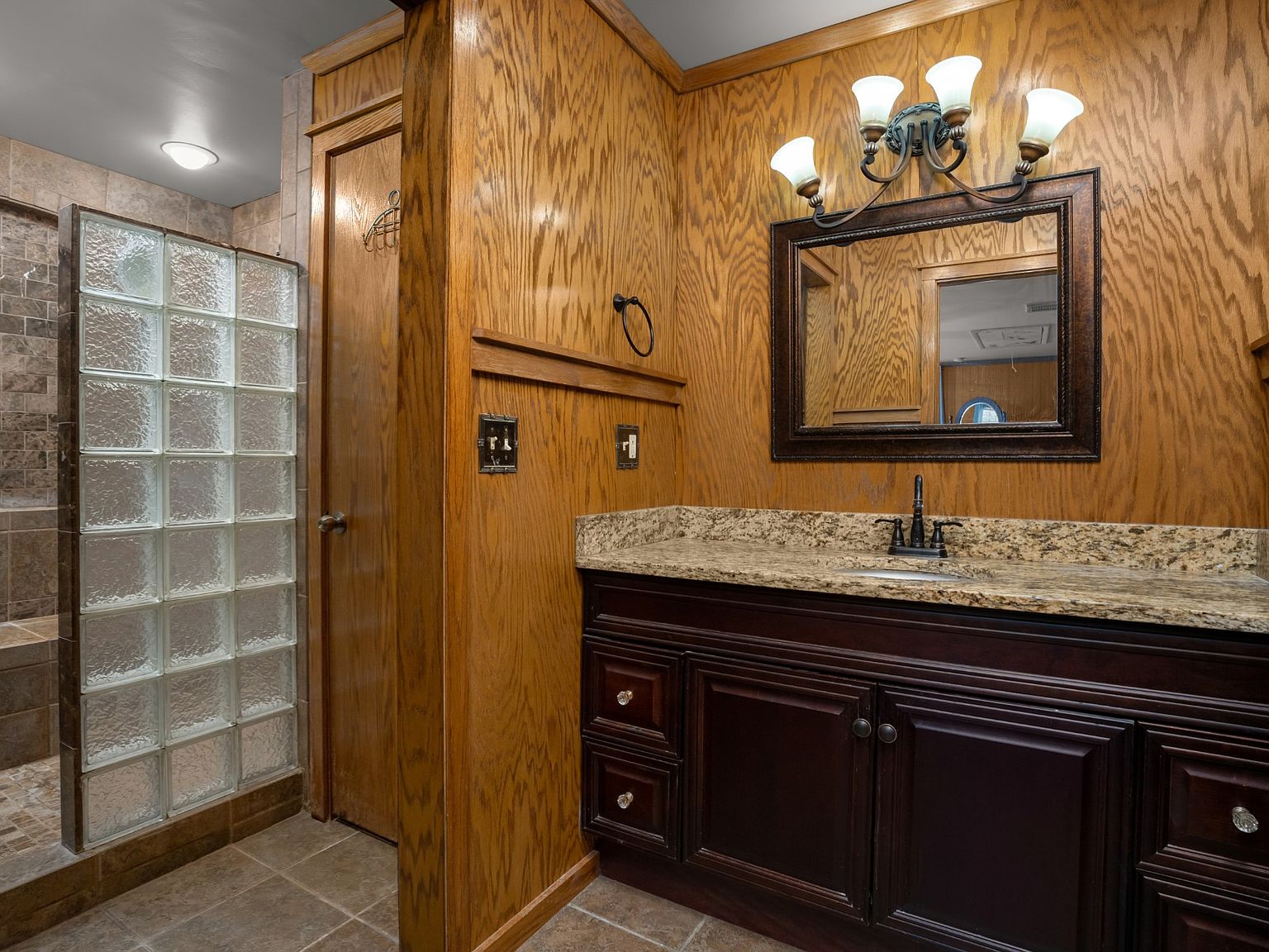 Bathroom with walnut wood cabinetry, granite countertop, and a glass-block shower nearby.