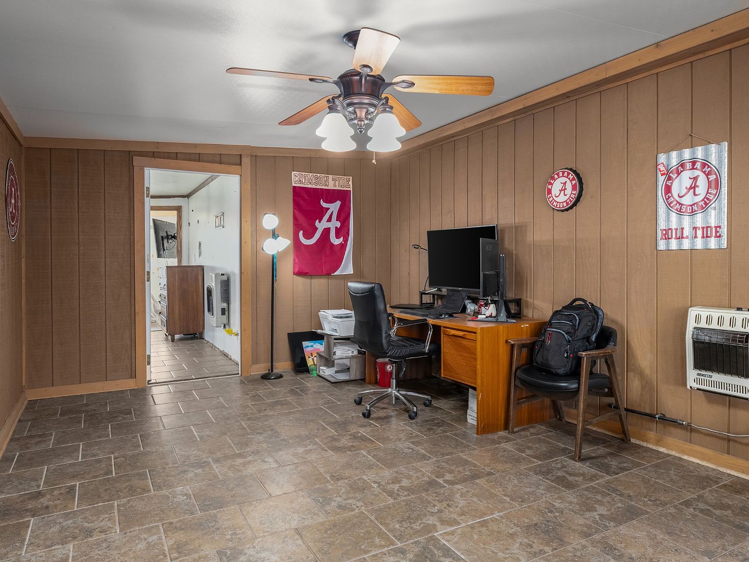 Home office in a wood-paneled room with Alabama Crimson Tide banners, a desk with dual monitors, and a black swivel chair.