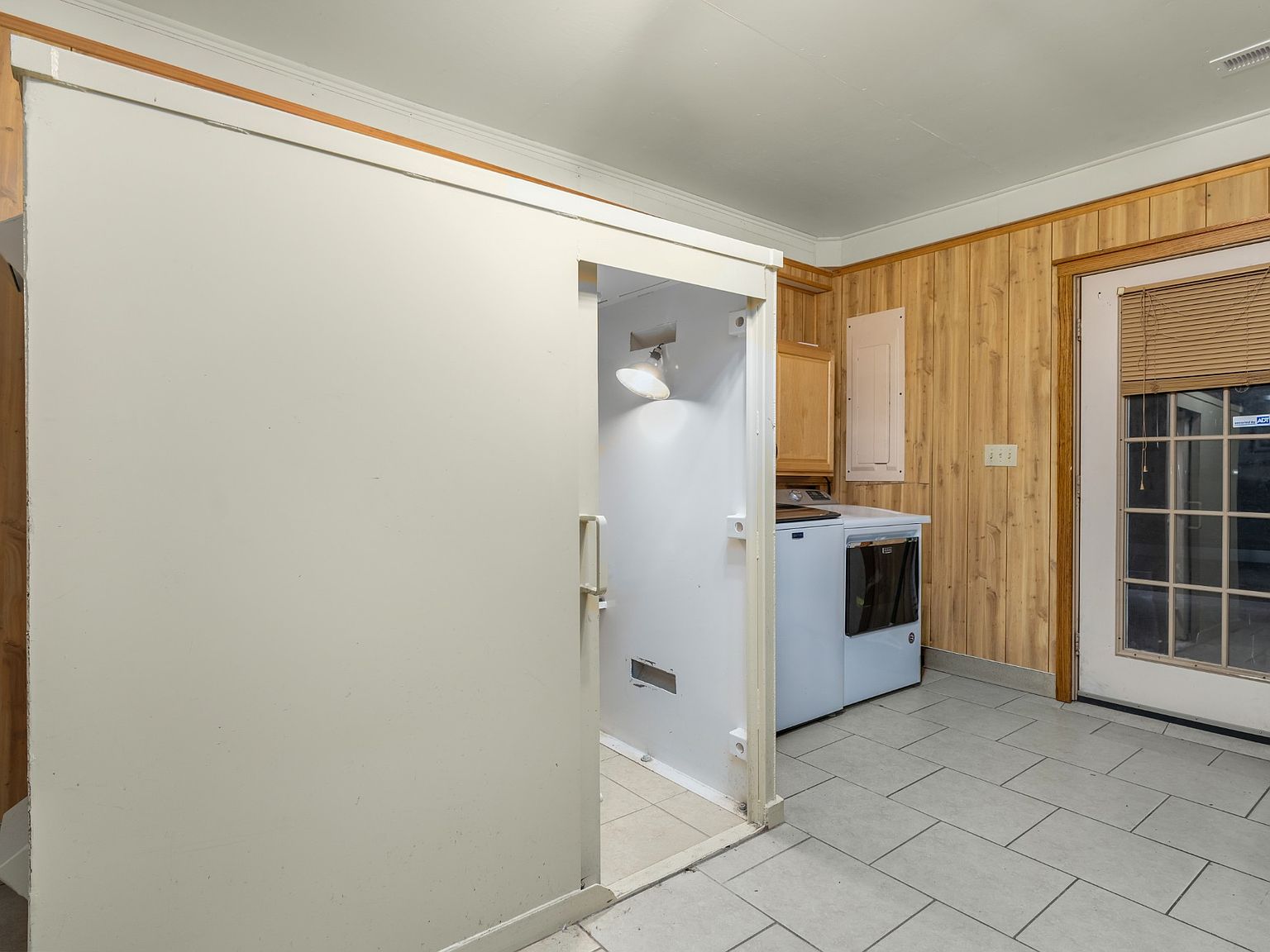 Laundry room with stacked washer and dryer beside a wood-paneled wall and a door with blinds; a white cabinet or closet is open in the foreground.