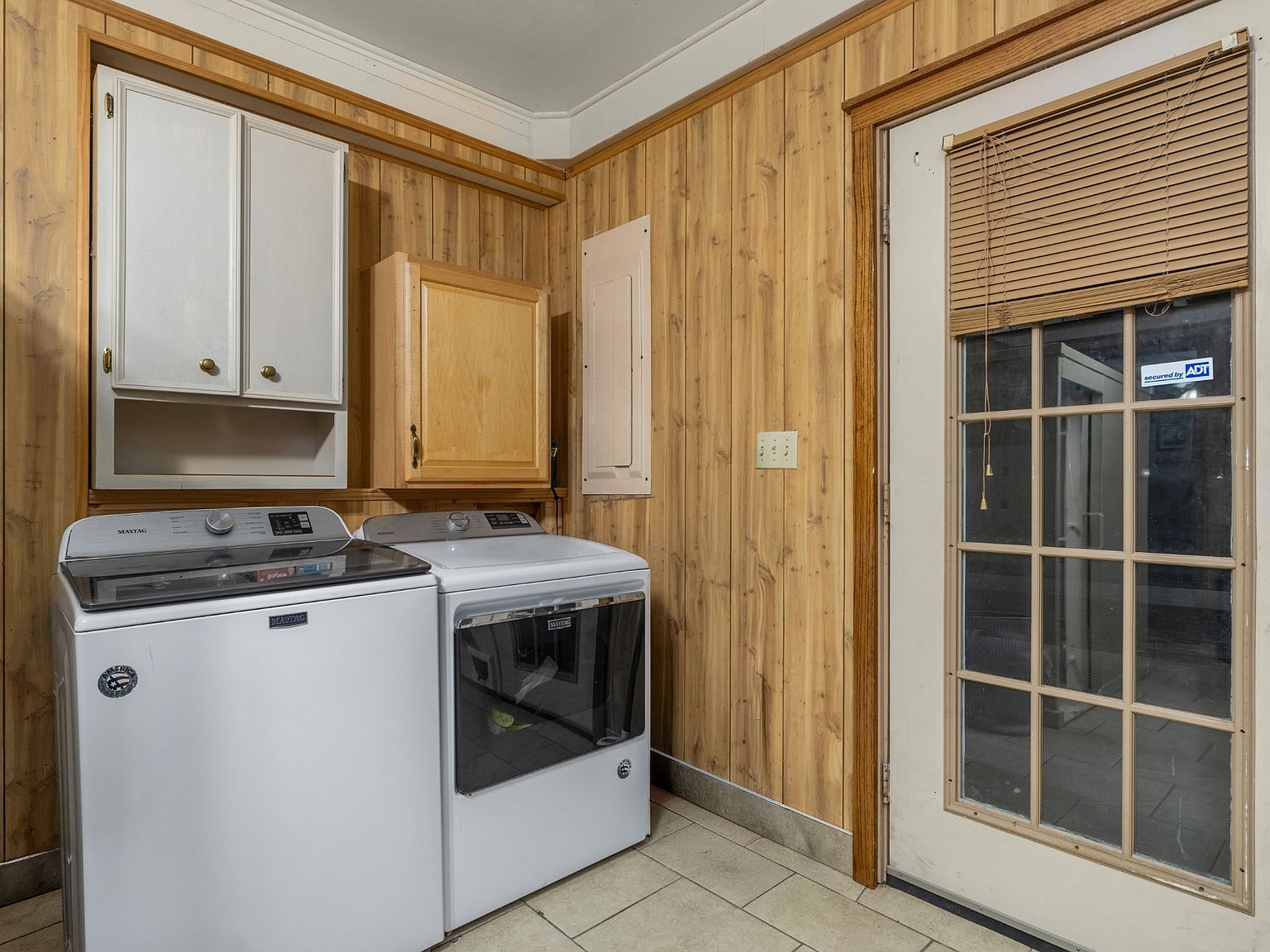Laundry room with white front-loading washer and dryer under light wood cabinets on paneled walls.
