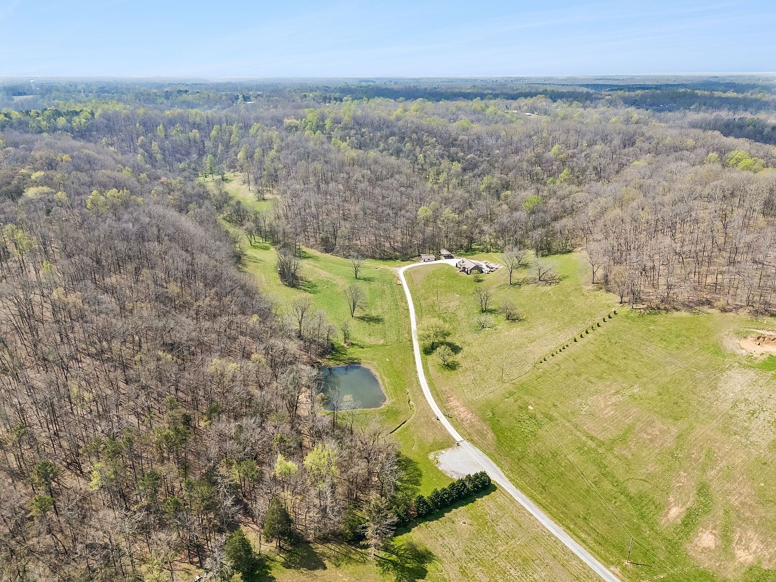 Aerial view of a rural property: a curved driveway leads to a house and outbuildings amid open green fields, with a pond to the left and dense woods surrounding the area.