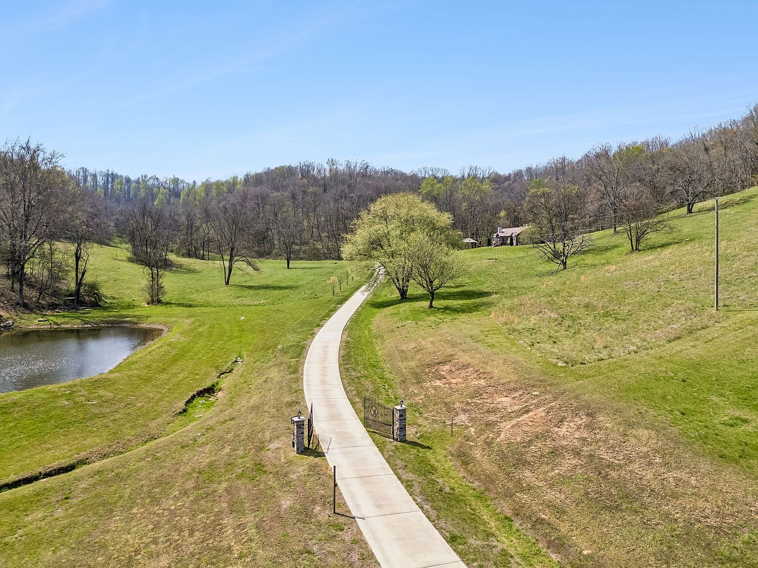 Wide view of a winding concrete path through a grassy countryside with a gate at the foreground and a pond to the left, trees dotting the hills.