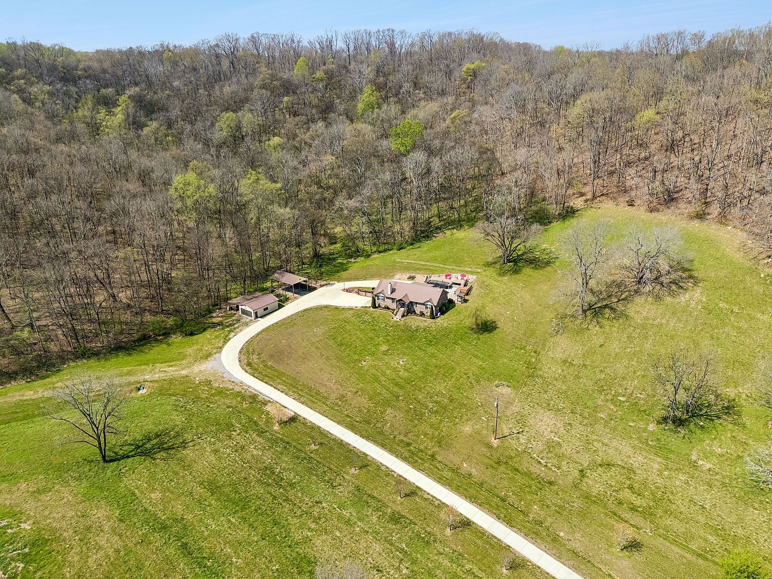 Aerial view of a single-story house with a curved driveway in a grassy clearing, bordered by dense leafless trees.