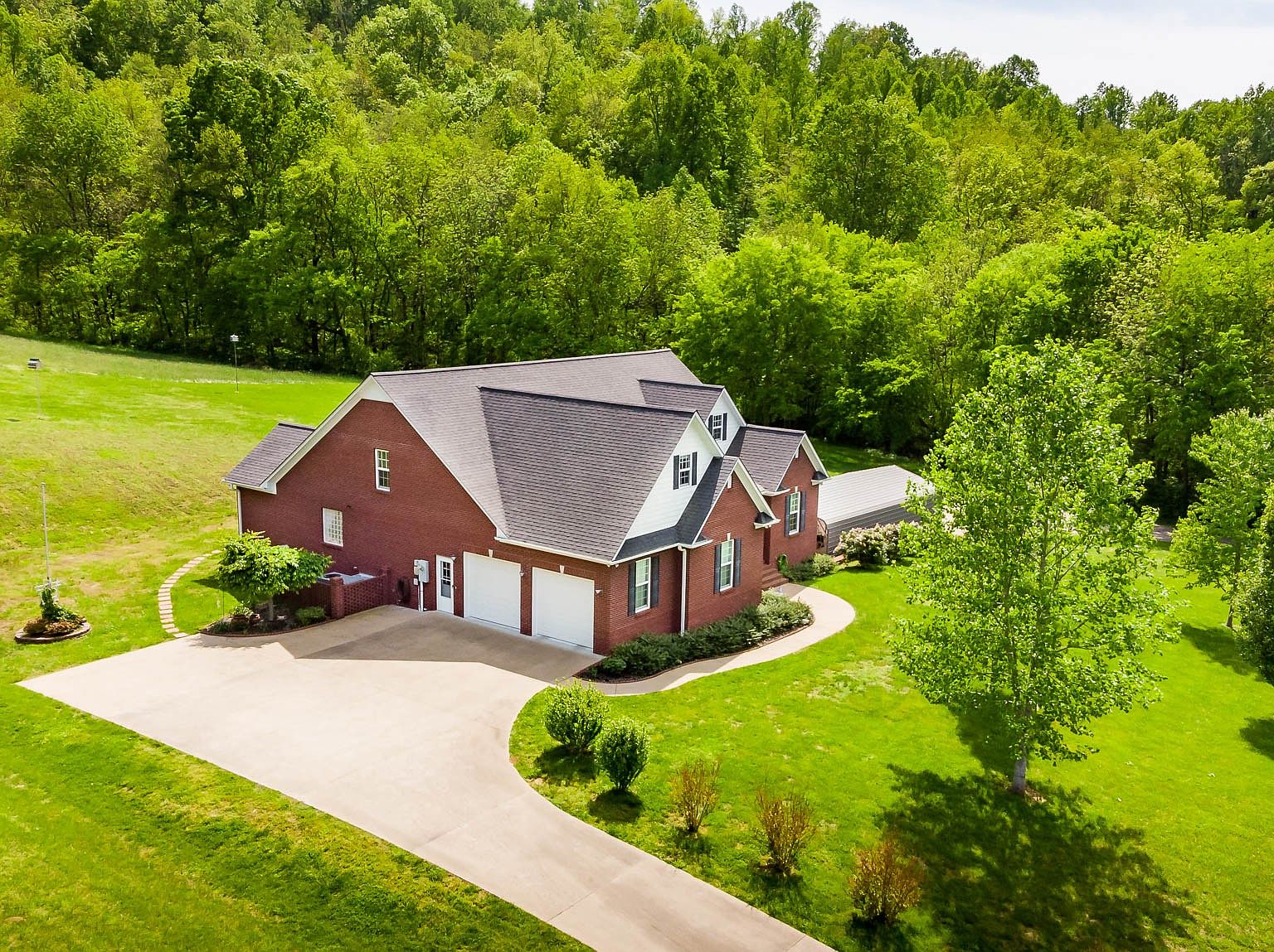 Brick house with a three-car garage, curved concrete driveway, and well-kept lawn bordered by trees and woods in the background.