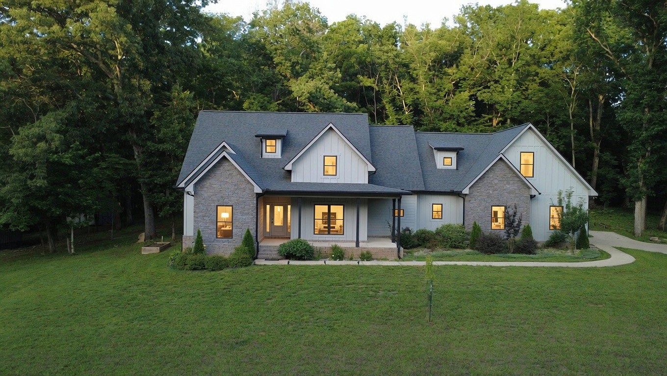 Two-story modern house with stone and light-gray siding, warm-lit windows, surrounded by trees and a curved driveway at dusk.