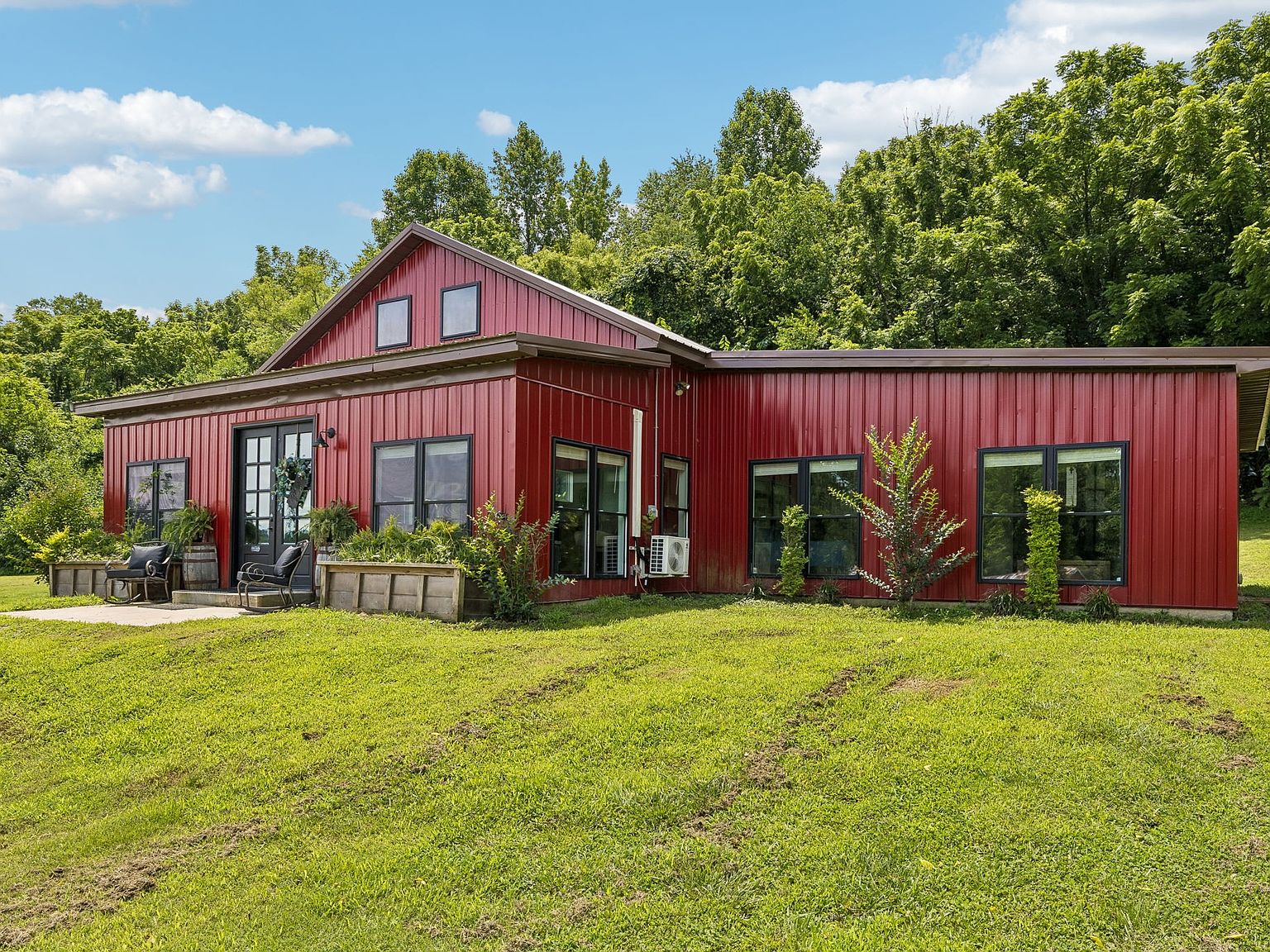Red metal-roofed house with large black-framed windows, a small porch, and planters in a grassy yard against green trees.