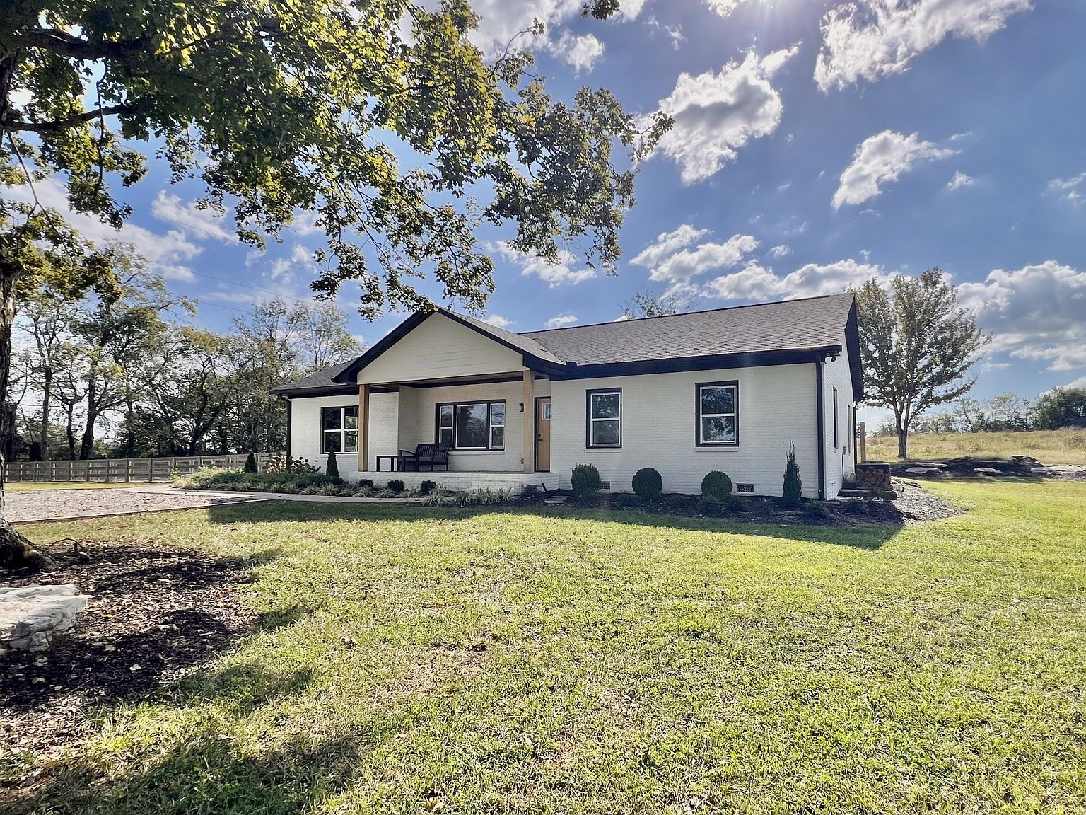 Single-story white brick house with a front porch and dark trim, set on a grassy yard under a blue sky with scattered clouds.