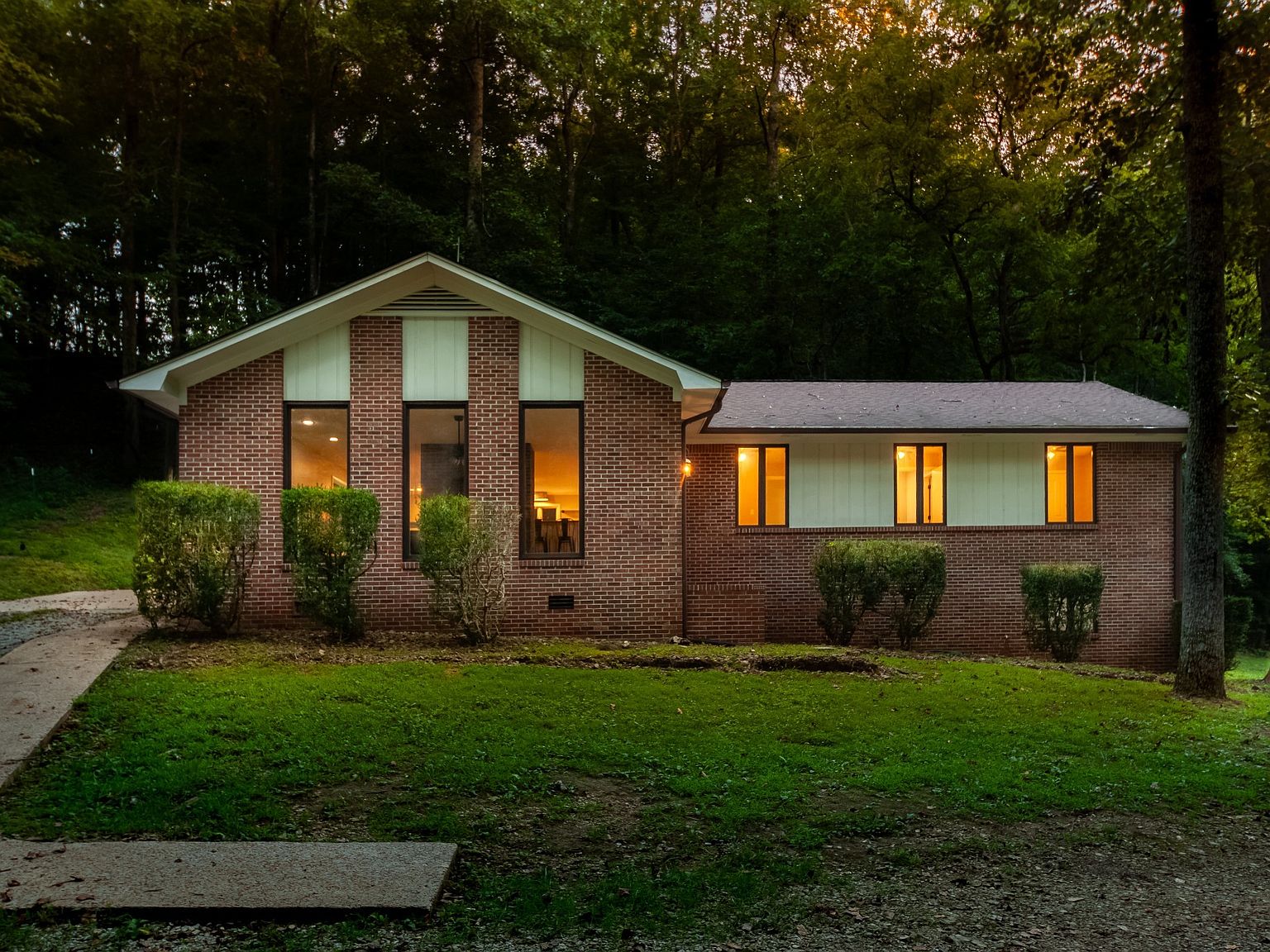 Brick, single-story house with large front windows and warm interior lights, set on a grassy lawn with neatly trimmed shrubs and a forest backdrop at dusk/early evening.