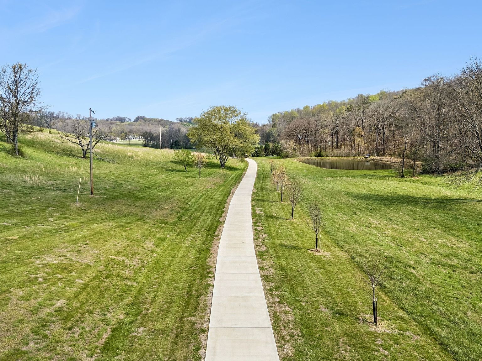 A straight concrete path runs through a green park, with a pond to the right and trees along rolling hills under a bright blue sky.