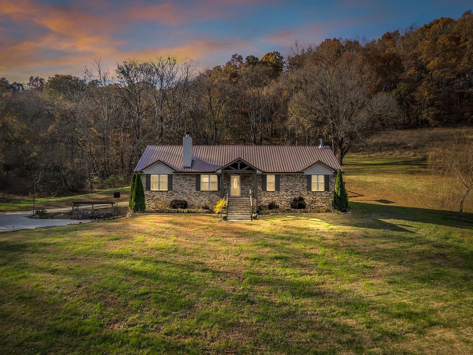Single-story brick house with a metal roof sits on a grassy clearing, with a forest backdrop at sunset.