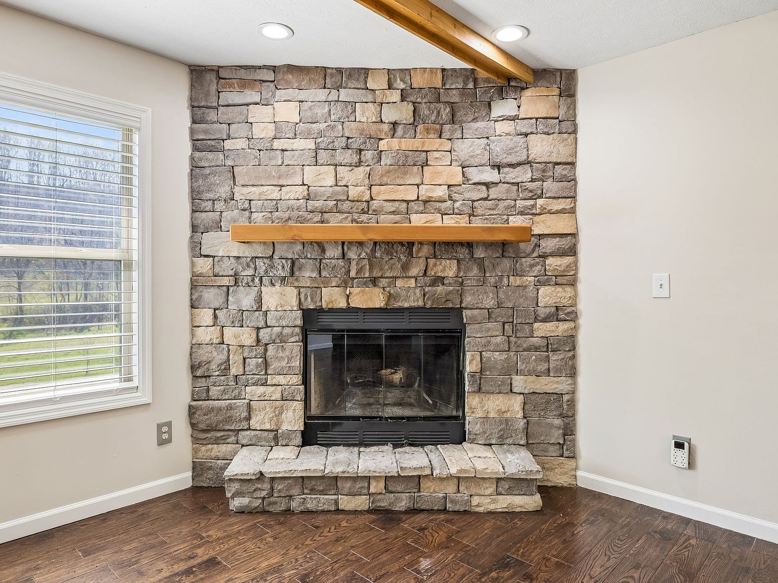 Stone fireplace wall with a wooden mantel and raised stone hearth in a living room, window to the left and wood floors nearby.