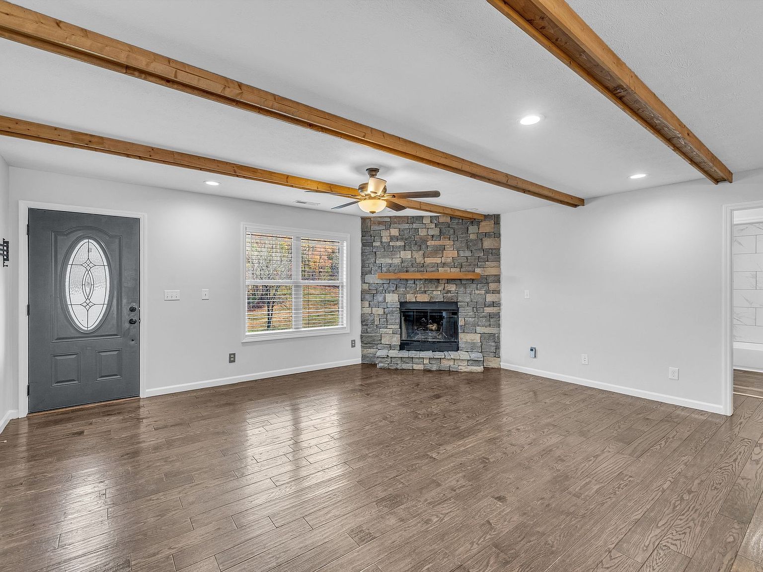 Living room with a stacked-stone fireplace, wood ceiling beams, and hardwood floors near a gray front door