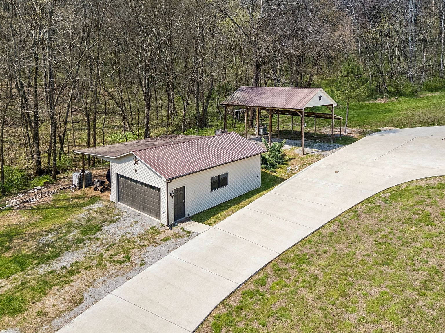 A small white garage with a brown metal roof and a large dark garage door, beside a curved concrete driveway. Behind it is a wooden open-sided shelter with a star. Trees border the property.