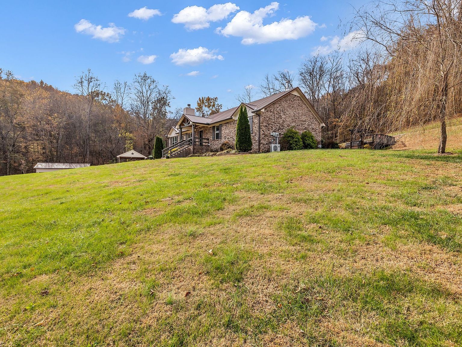 Stone cottage with a metal roof on a grassy hill, surrounded by trees under a blue sky.