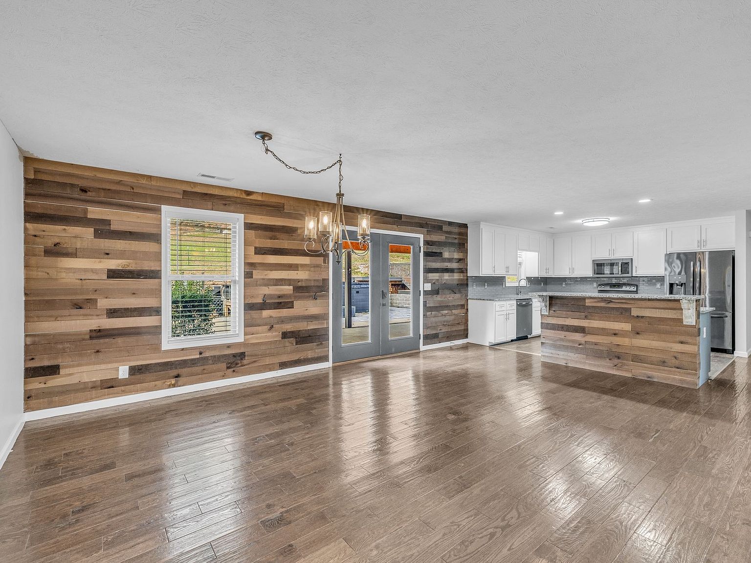 Open-concept living space with a rustic wood plank accent wall, large window, French doors, and a white kitchen with an island.
