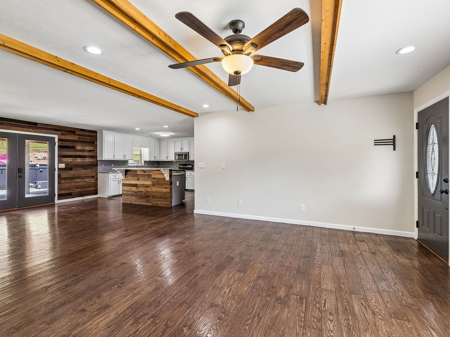 Open living area with dark hardwood floors, exposed wooden ceiling beams, and a ceiling fan; kitchen with white cabinets and a wood-paneled island visible in the background.