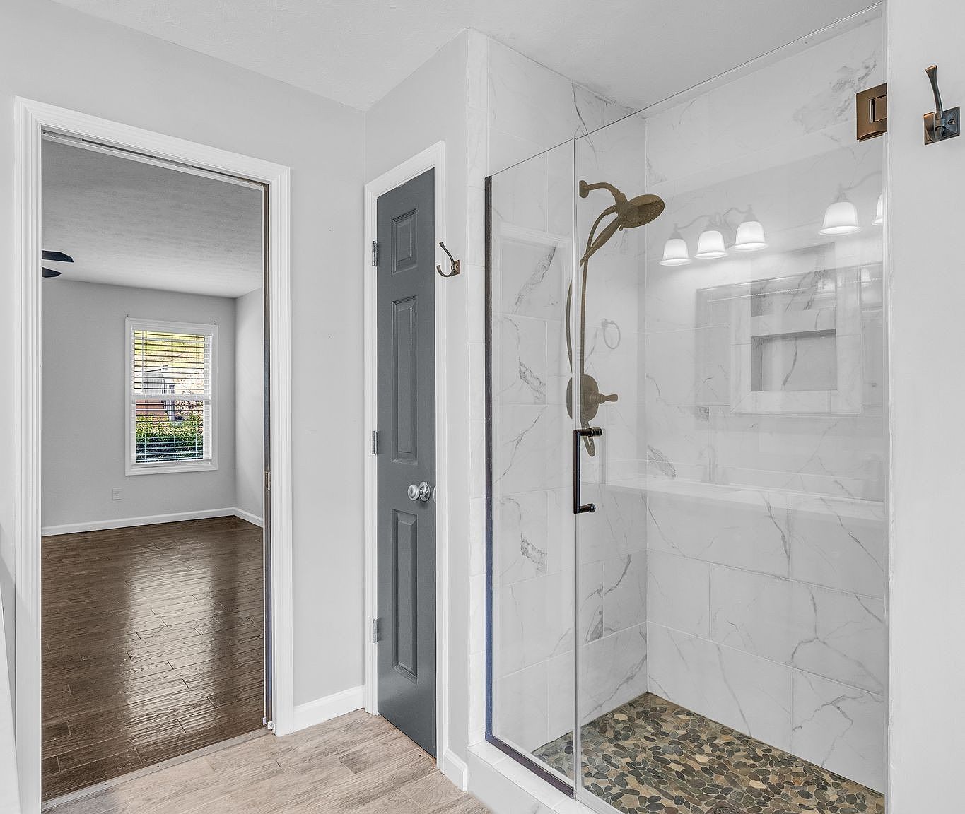Glass-walled shower with marble walls and a pebble floor, next to a gray interior door in a bright bathroom. Nearby a doorway leads to a room with wood flooring and a window.