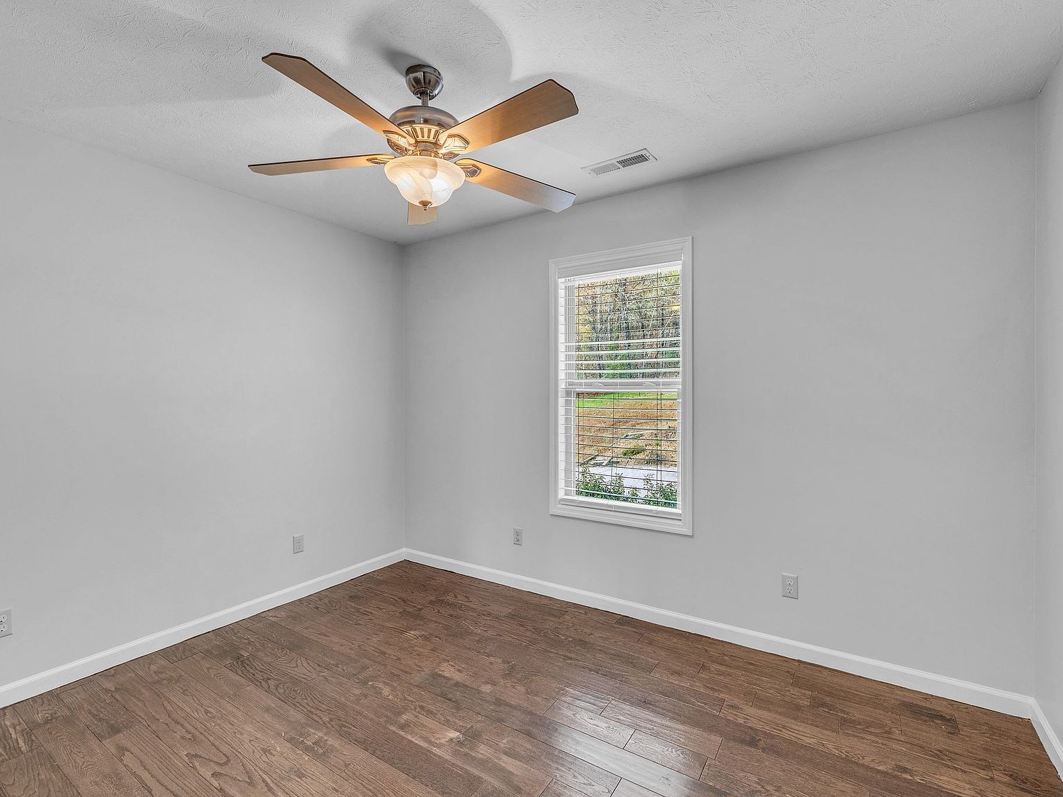 Empty bedroom with gray walls, dark wooden floors, a white baseboard, window with blinds, and a ceiling fan/light fixture.